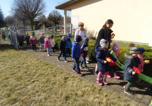 Dzieci wraz z panią dyrektor maszerują z wiosennymi plakatami, panią Wiosną pod budynkiem przedszkola.